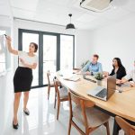 Young business trainer pointing at whiteboard and conducting business training for business people who sitting at the table and listening to her at office